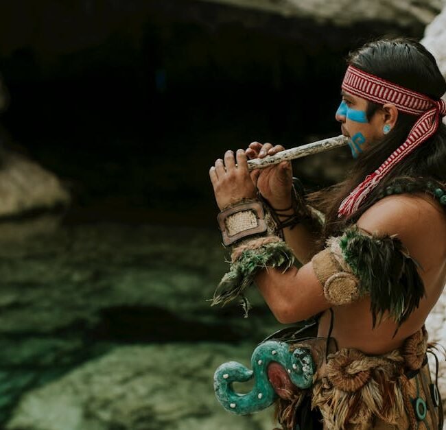 A Mayan musician wearing traditional attire plays a flute by a rocky water landscape, exuding cultural heritage and art.